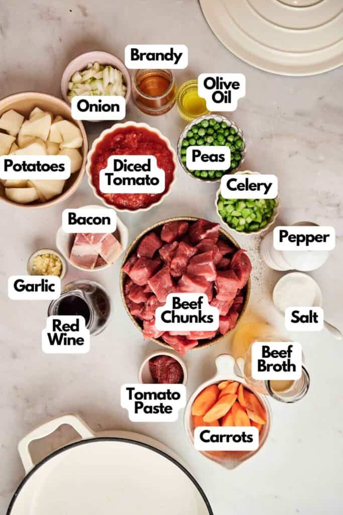 Ingredients for a beef stew laid out on a countertop, including beef chunks, potatoes, carrots, peas, diced tomato, celery, bacon, and various seasonings and liquids.