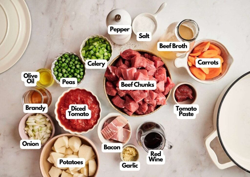 Ingredients for a hearty Beef Stew arranged on a table, featuring beef chunks, potatoes, carrots, peas, celery, onion, bacon, garlic, diced tomato, tomato paste, beef broth, red wine, brandy, olive oil, salt and pepper.