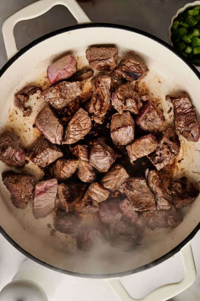 Sautéing cubed beef pieces in a white pan, with steam rising and diced green bell peppers in a small bowl nearby.