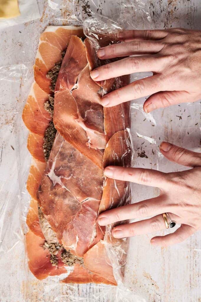 Hands are wrapping prosciutto around a savory filling for a beef wellington, using plastic wrap on a wooden surface.