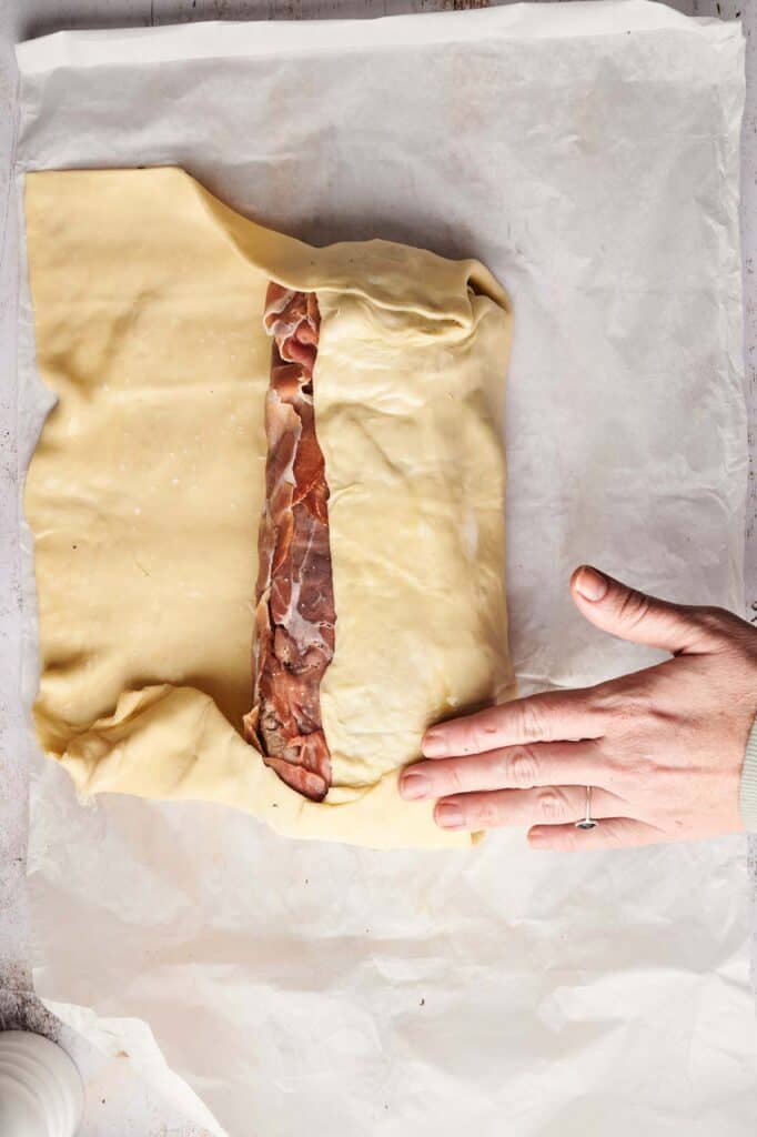 A hand prepares a pastry akin to beef Wellington, carefully layering thin slices of meat on parchment paper.