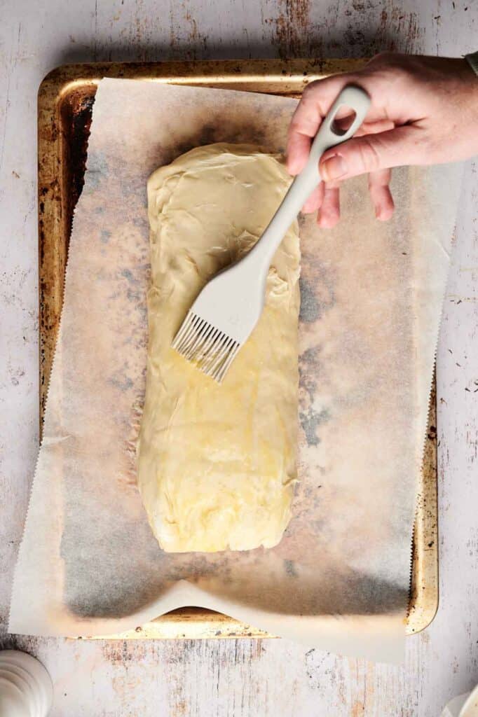A hand brushes a pastry with melted butter on a parchment-lined baking sheet, prepping the perfect crust for a delectable beef wellington.