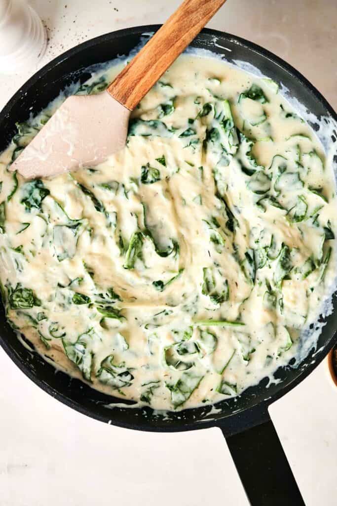 A pan filled with creamy spinach dip, reminiscent of a luscious Chicken Alfredo Lasagna, is being stirred gently with a spatula.