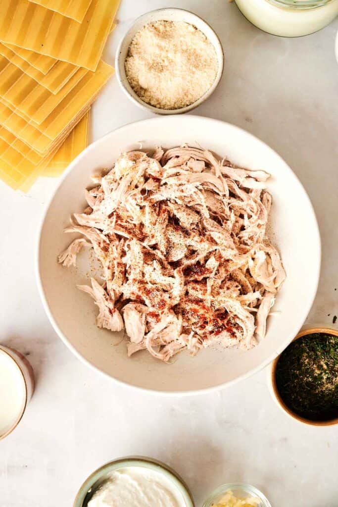 A bowl of shredded chicken, seasoned to perfection, is surrounded by lasagna sheets and grated cheese, a delightful twist for a Chicken Alfredo Lasagna masterpiece. Fresh herbs generously garnish the marble surface, promising an elegant feast with every bite.