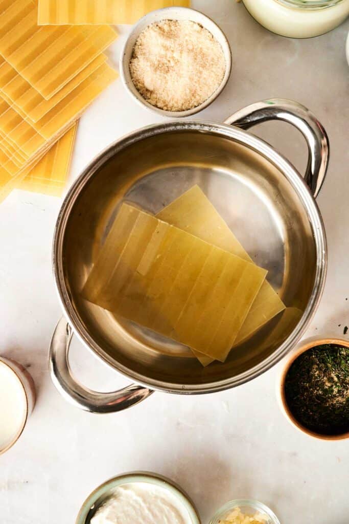 Top view of uncooked chicken Alfredo lasagna sheets in a metal pot, surrounded by bowls of ingredients like cheese, sauce, and herbs on a white surface.
