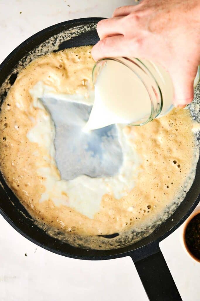 A hand pours milk from a glass jar into a frying pan containing a bubbling mixture reminiscent of Chicken Alfredo Lasagna.
