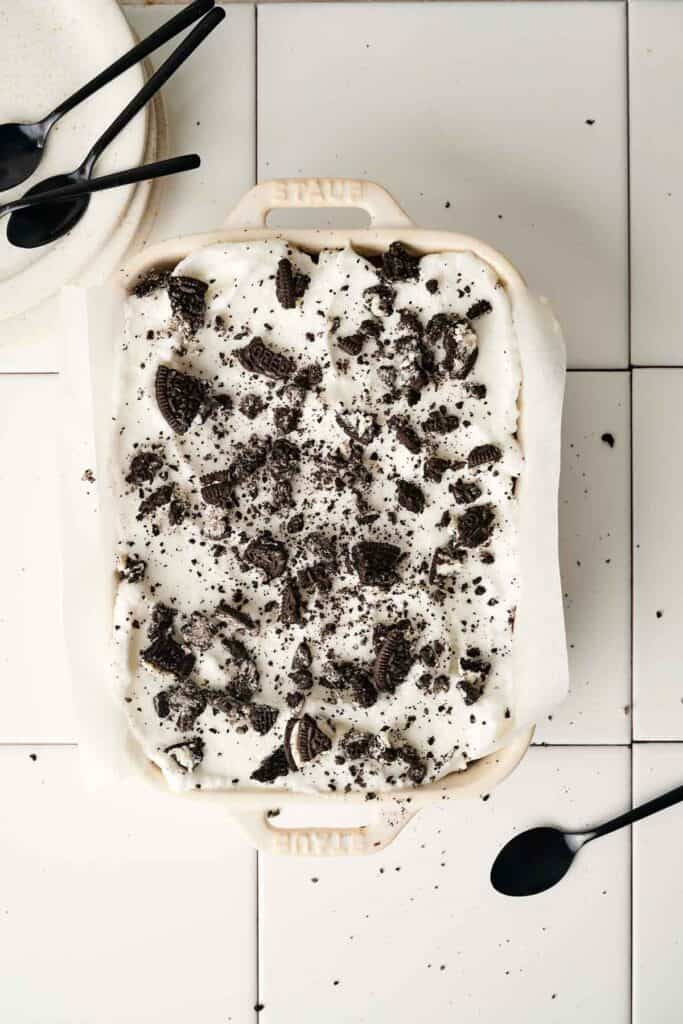 A delightful dish of chocolate lasagna dessert with whipped topping and crushed cookies sits on a tiled surface. Nearby, a stack of plates and spoons awaits eager guests.