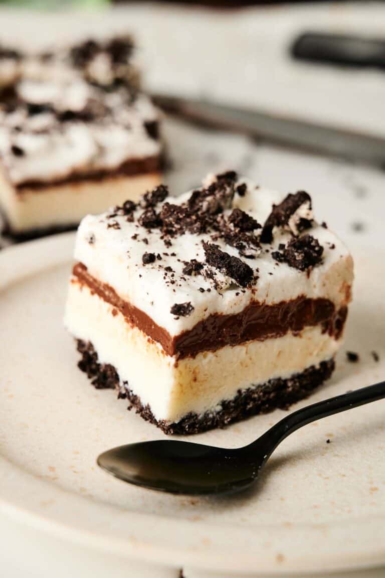A slice of chocolate lasagna dessert with a cookie crust, creamy filling, rich chocolate layer, and whipped topping, garnished with crushed cookies, sits invitingly on a plate accompanied by a spoon.