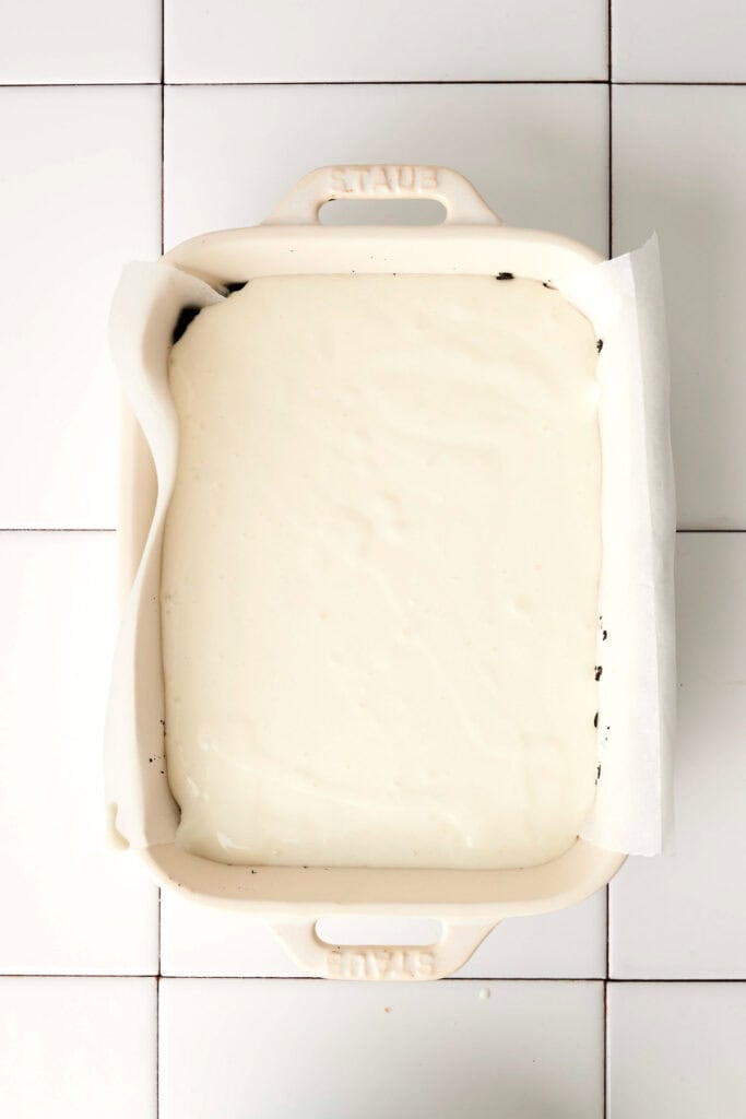 A rectangular baking dish lined with parchment paper holds a creamy white batter, reminiscent of a luscious chocolate lasagna, set gracefully on a white tiled surface.