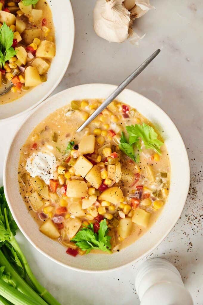 A bowl of corn chowder with potatoes, visible seasonings, and garnished with fresh herbs. Corn adds a sweet crunch to this creamy delight. A spoon rests in the bowl, ready to dig in.