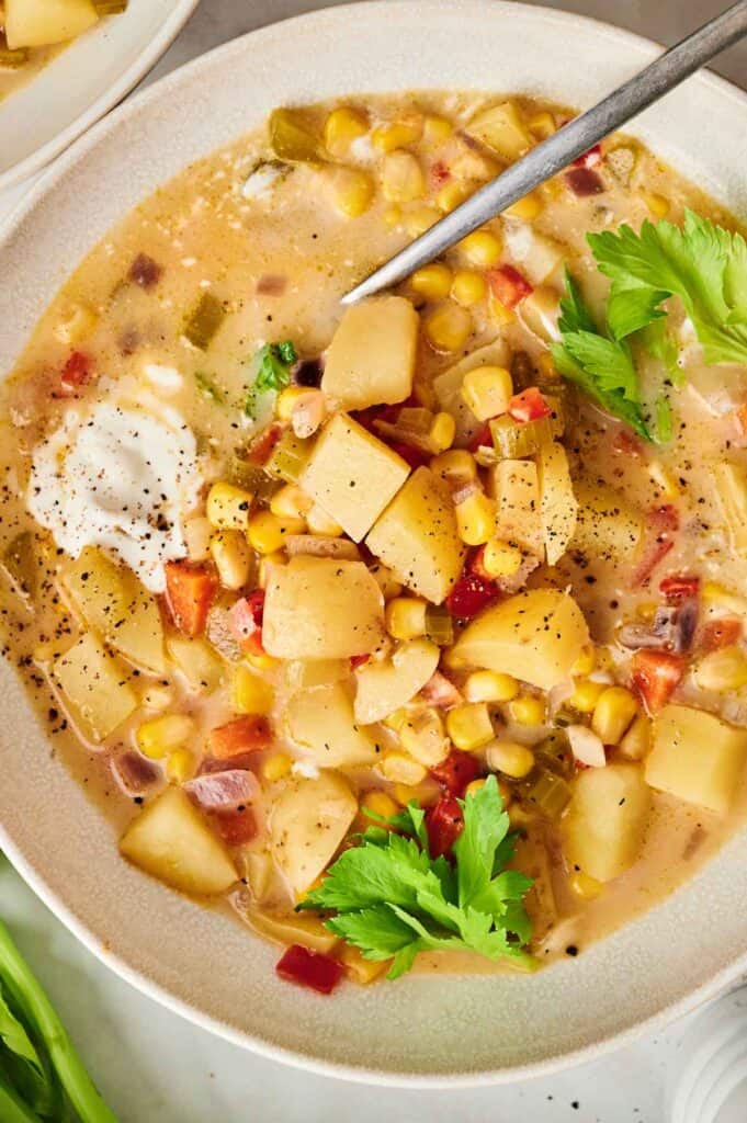 A bowl of creamy Corn Chowder brimming with potatoes, red peppers, and fresh herbs, garnished with parsley. A spoon rests gracefully in the rich, flavorful mixture.