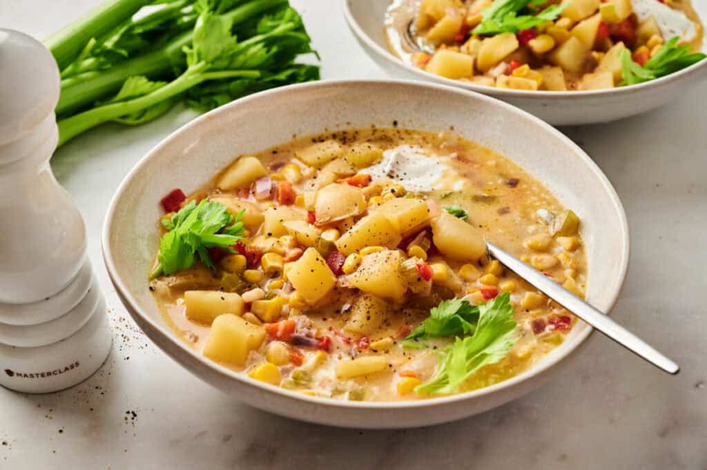 A bowl of hearty corn chowder with potatoes, corn, and herbs, garnished with celery leaves. A spoon rests in the bowl, and celery stalks and a pepper grinder are visible in the background.