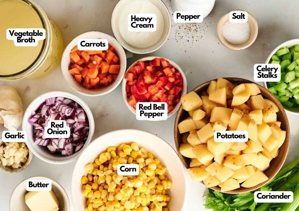 Ingredients for a Corn Chowder soup are displayed on a counter, including chopped potatoes, carrots, celery, red onion, corn, red bell pepper, garlic, vegetable broth, butter, heavy cream, salt, pepper, and coriander.