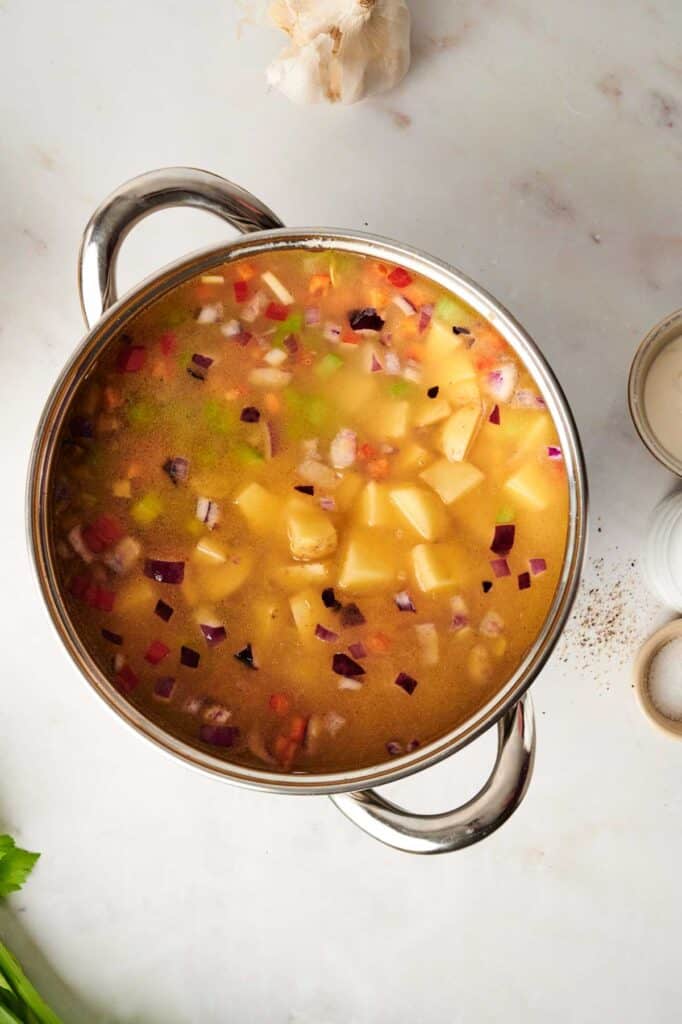A pot of hearty vegetable soup, reminiscent of a comforting corn chowder, features diced potatoes, onions, and other vegetables on a marble countertop, surrounded by garlic and seasonings.