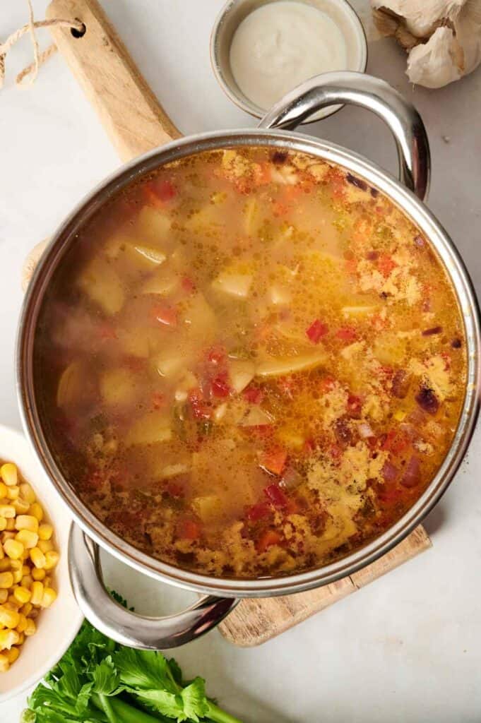 A pot of vegetable soup with visible chunks of potatoes, tomatoes, and spices sits on a wooden board. Nearby are a bowl of corn reminiscent of corn chowder, a small dish of sour cream, garlic, and fresh herbs.