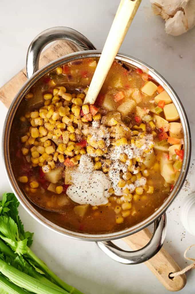 A pot of delicious corn chowder brimming with diced vegetables, topped with black pepper and cream, sits invitingly with a wooden spoon inside; fresh celery and garlic rest nearby on a cutting board.