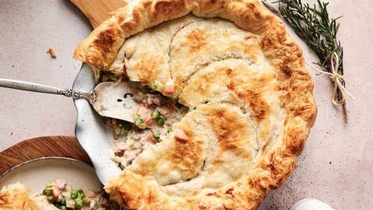 A golden-brown chicken pot pie with a slice being served, showing chunks of chicken, peas, and carrots. A sprig of rosemary is nearby.