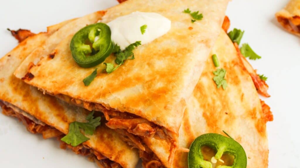 Quesadillas stacked on a plate, topped with sliced jalape&ntilde;os, cilantro, and a dollop of sour cream.