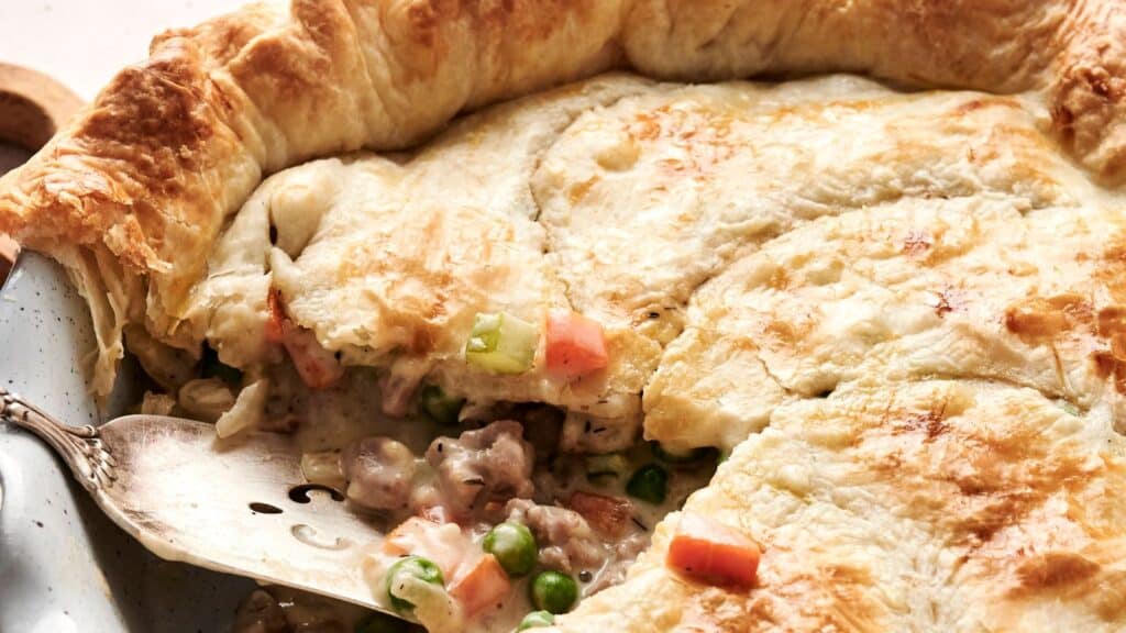 A golden-brown pot pie with a flaky crust is partially sliced, revealing a filling of peas, carrots, celery, and meat on a serving spatula.