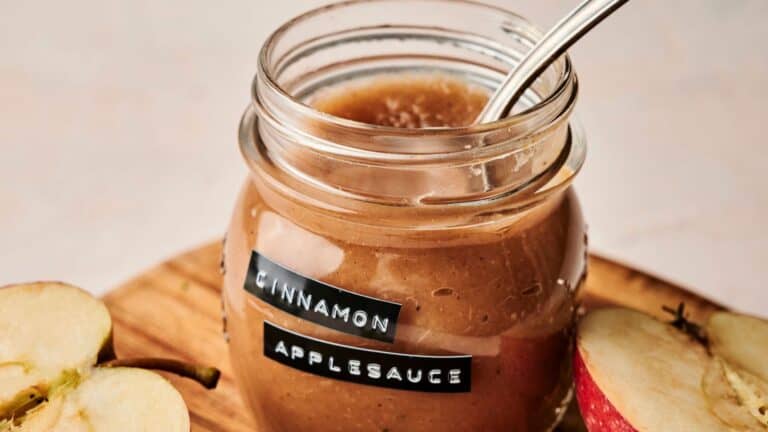 A jar of cinnamon applesauce with a metal spoon inside. The jar is on a wooden surface next to sliced apples.
