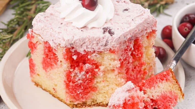 A slice of cake with red swirls and pink frosting topped with whipped cream and a cherry sits on a plate. A fork holds a piece of cake.