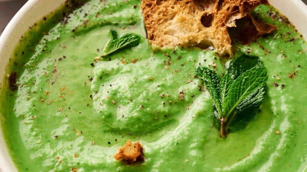A bowl of creamy green pea soup garnished with mint leaves, black pepper, and pieces of crusty bread.