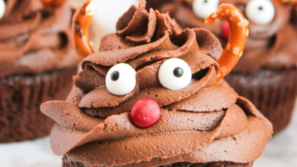 Chocolate cupcake with swirled frosting, candy eyes, a red candy nose, and pretzel antlers, resembling a reindeer.