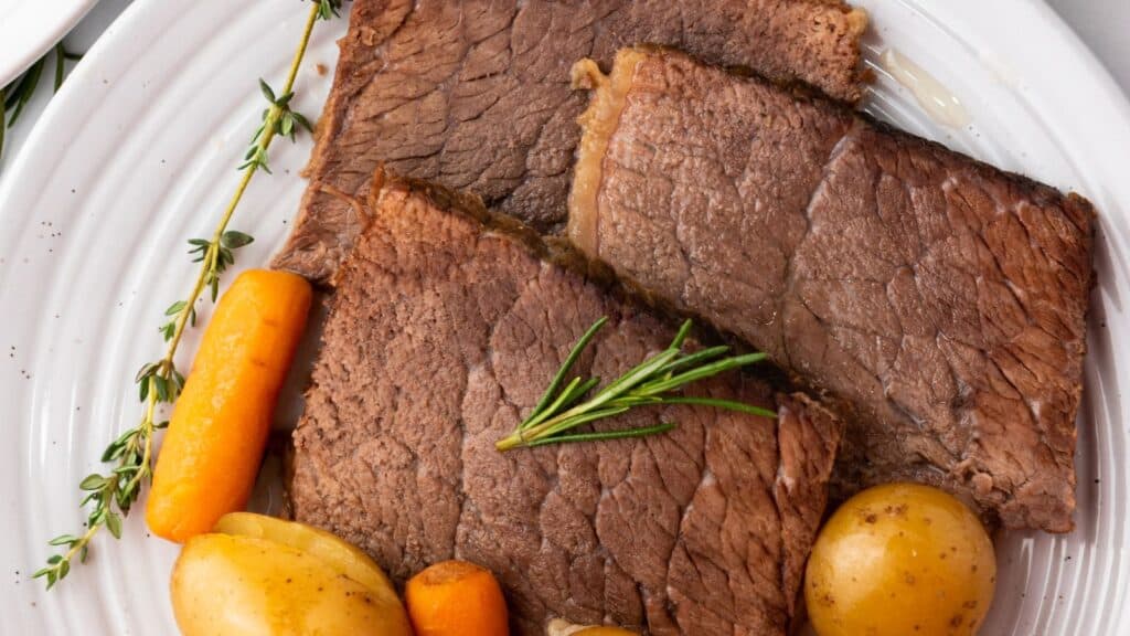 Slices of cooked roast beef with rosemary on top, accompanied by carrots and potatoes on a white plate.