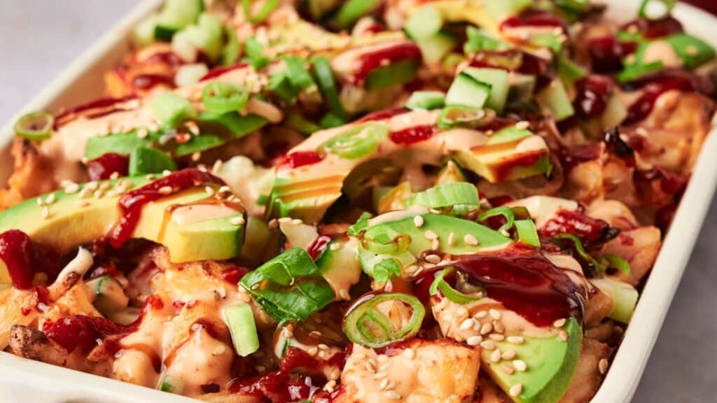 A dish of loaded fries topped with sliced avocado, green onions, cucumbers, sesame seeds, and drizzles of creamy and red sauces.