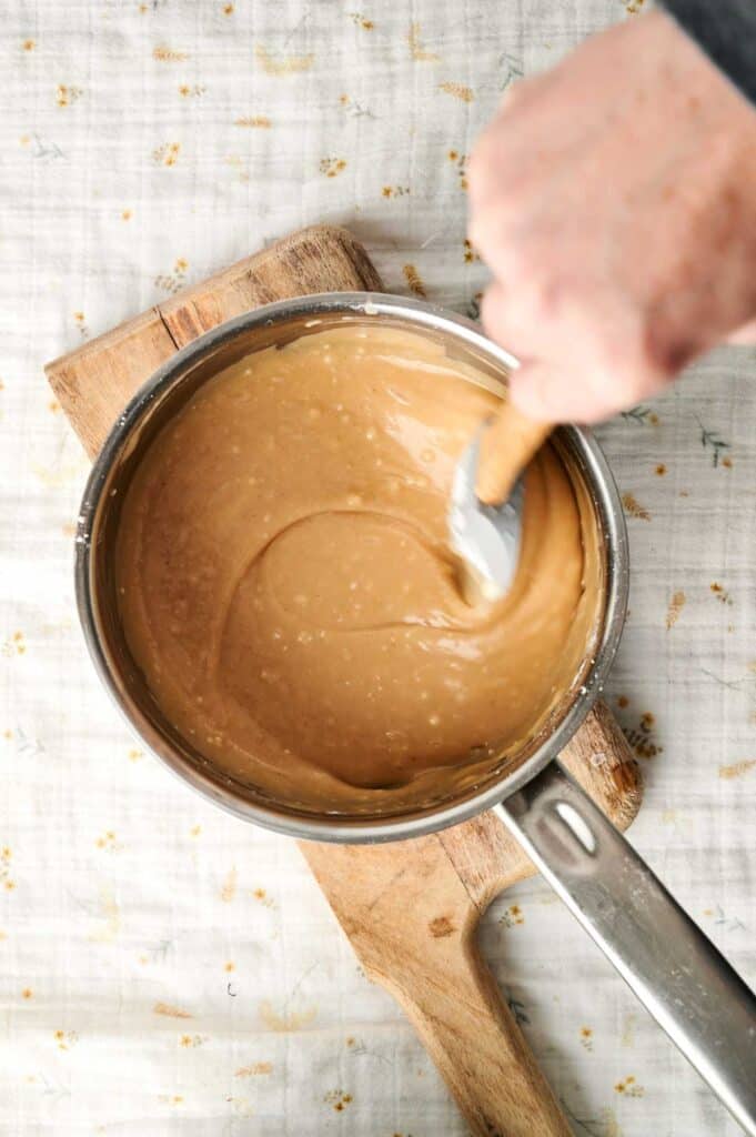 A hand expertly stirs peanut butter mixture in a metal saucepan with a spatula on a wooden board, preparing the perfect topping for a rich Peanut Butter Cake.