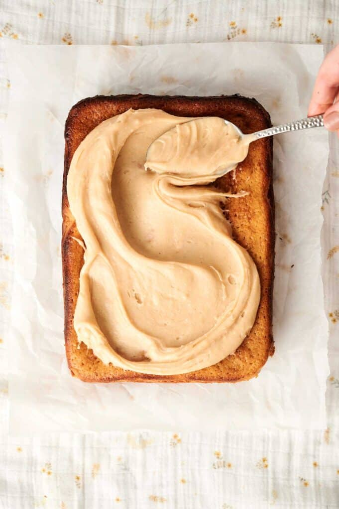 A rectangular peanut butter cake is being frosted with thick, creamy brown icing using a spoon. The cake sits on parchment paper.