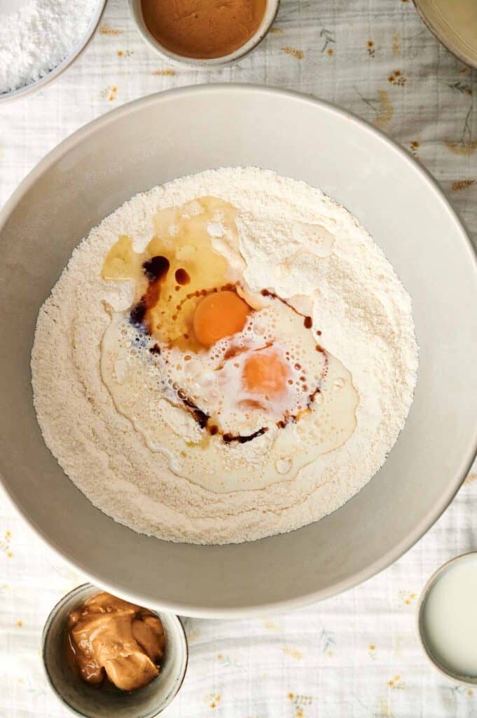 A mixing bowl with flour, an egg, and liquid ingredients sits ready. Nearby, small bowls of peanut butter and other essentials for crafting a delectable peanut butter cake rest on a patterned cloth.