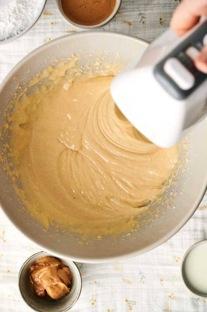 An electric mixer whirs smoothly, expertly blending batter in a large bowl atop a tablecloth, encircled by bowls of ingredients for a delectable Peanut Butter Cake, including creamy peanut butter and fresh milk.