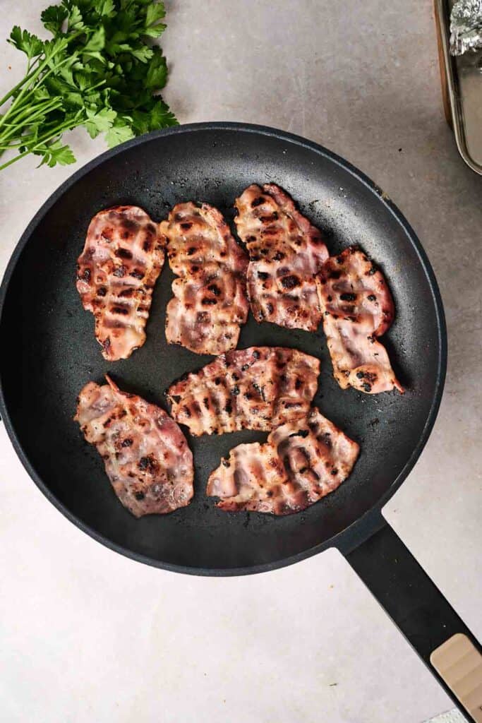 Slices of bacon sizzle in a black frying pan on a light-colored countertop, with fresh parsley nearby, ready to be sprinkled over a hearty bowl of potato soup.
