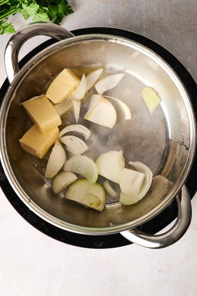 A pot on a stovetop contains sliced onions and chunks of butter, the perfect start for a savory potato soup. Parsley is in the top corner.