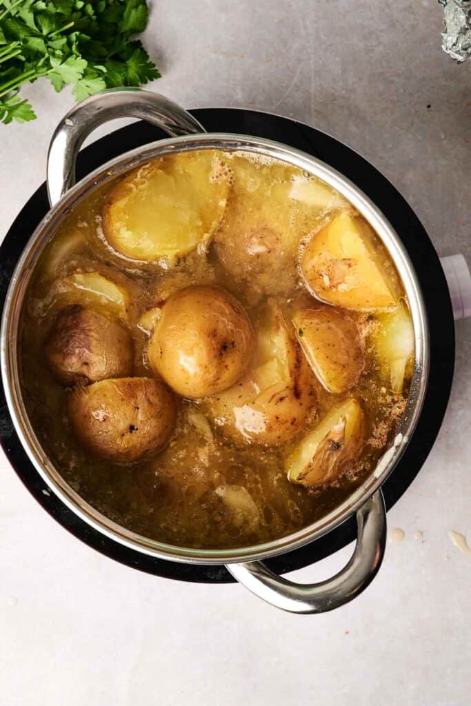 Potatoes destined for a comforting potato soup are boiling in a silver pot filled with water on the stovetop, with fresh herbs nearby to enhance the flavor.