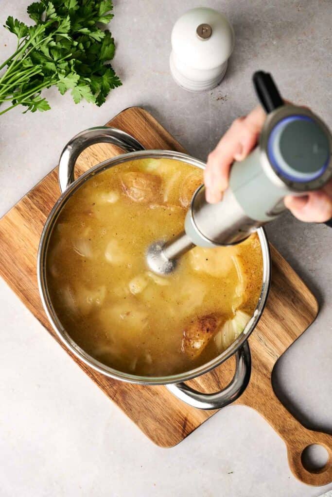 A hand uses an immersion blender in a pot of creamy potato soup on a wooden cutting board. Fresh parsley and a salt shaker are nearby.