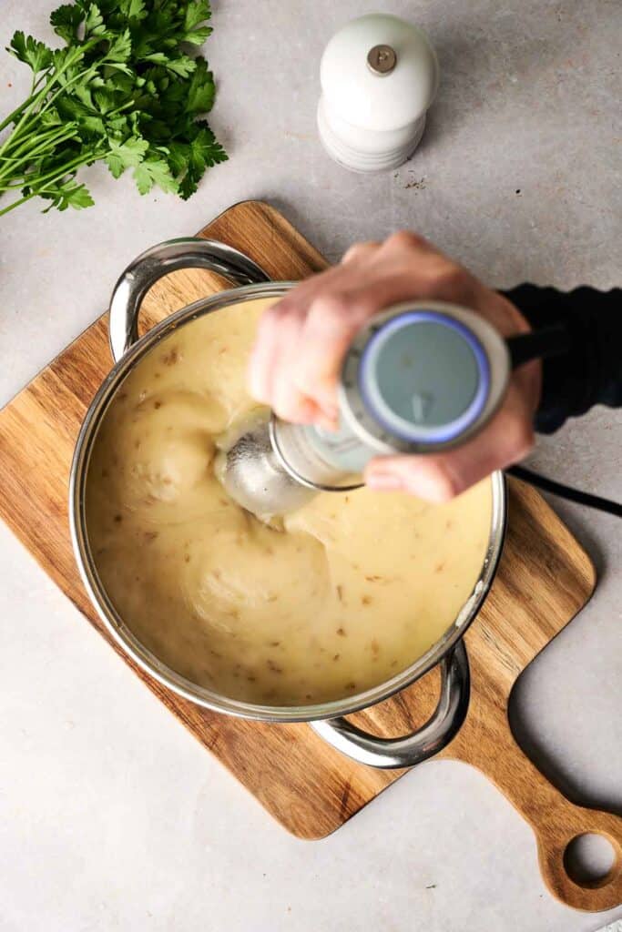 A person uses an immersion blender to puree creamy potato soup in a pot resting on a wooden board, surrounded by fresh herbs and a pepper grinder.