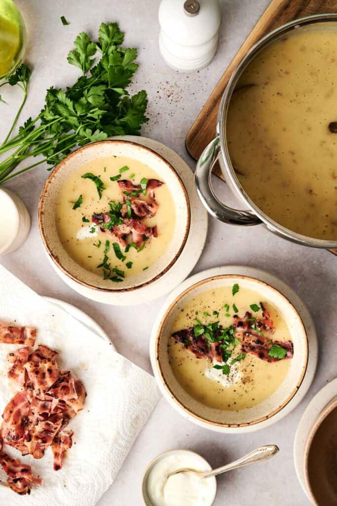 Two bowls of creamy potato soup are garnished with crispy bacon and herbs. Nearby are a pot of soup, crispy bacon on paper towels, herbs, and a small bowl of cream.