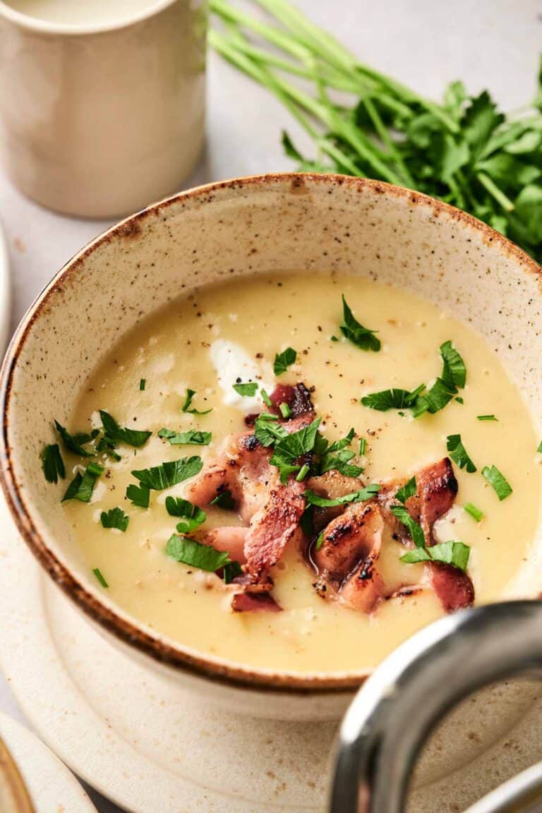 A bowl of creamy potato soup topped with crispy bacon pieces and chopped parsley, with a fresh parsley bunch in the background.