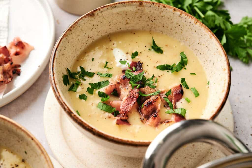 A bowl of creamy potato soup topped with chopped bacon and fresh parsley. A piece of bacon rests on a nearby plate, while parsley leaves add a touch of green in the background.