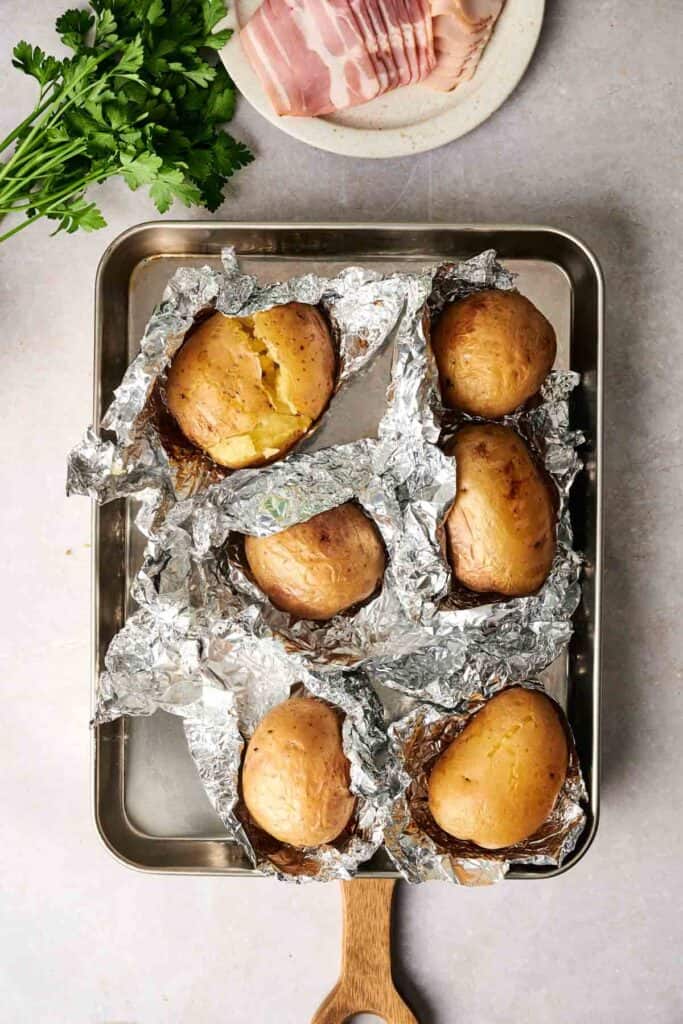 Six baked potatoes wrapped in foil rest on a baking tray, ready to be transformed into creamy potato soup. Nearby, bacon slices and fresh parsley await their turn to add flavor and flair.