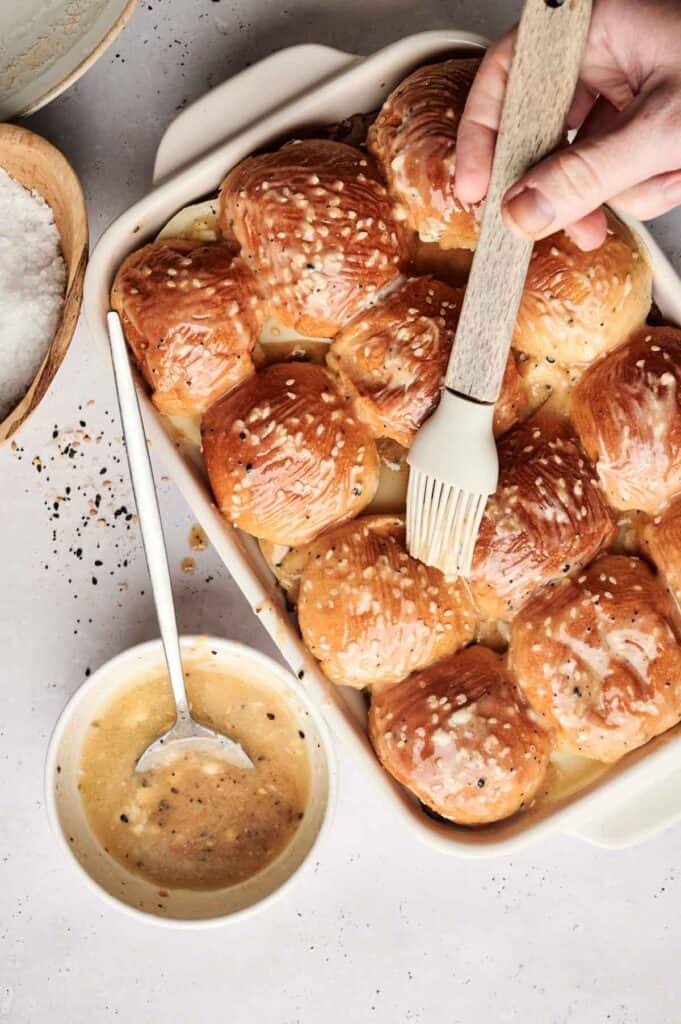 A hand brushes melted butter over pull-apart rolls in a baking dish, perfect for roast beef sliders. A bowl of butter with a spoon is nearby.