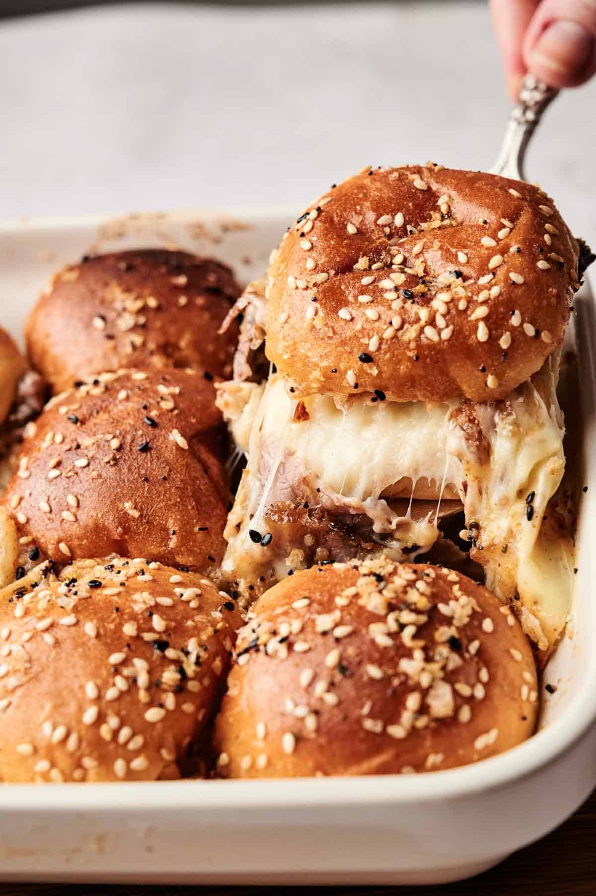 A tray of homemade cheeseburger sliders with sesame seed buns boasts a mouthwatering display; one slider is being lifted to reveal gooey, melted cheese, reminiscent of the savory goodness found in classic roast beef sliders.