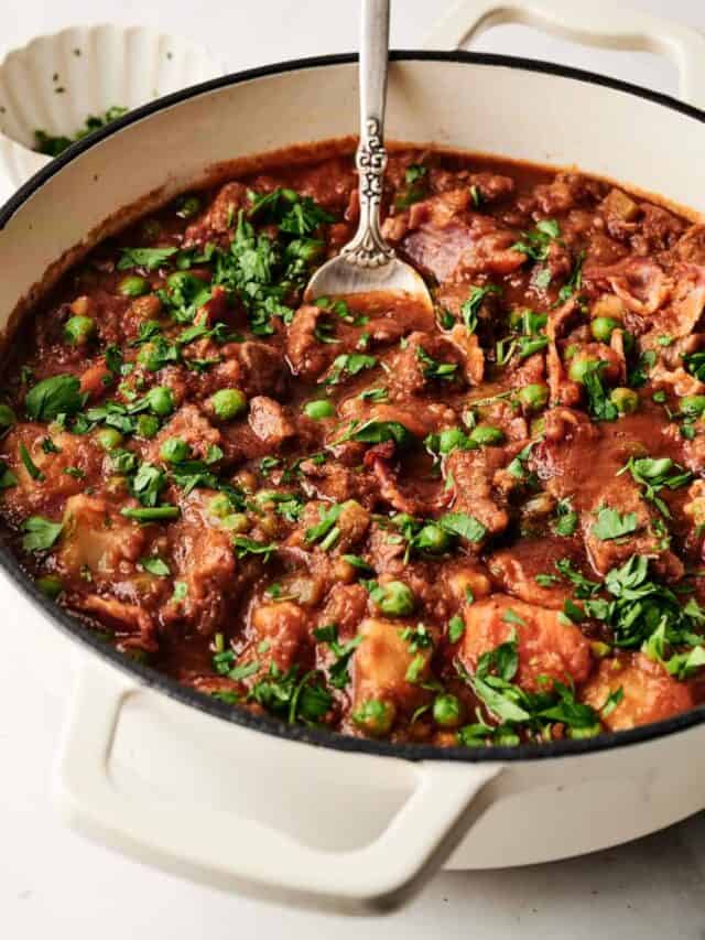 Hearty stew with meat, peas, and herbs in a white pot with a spoon on a white surface.