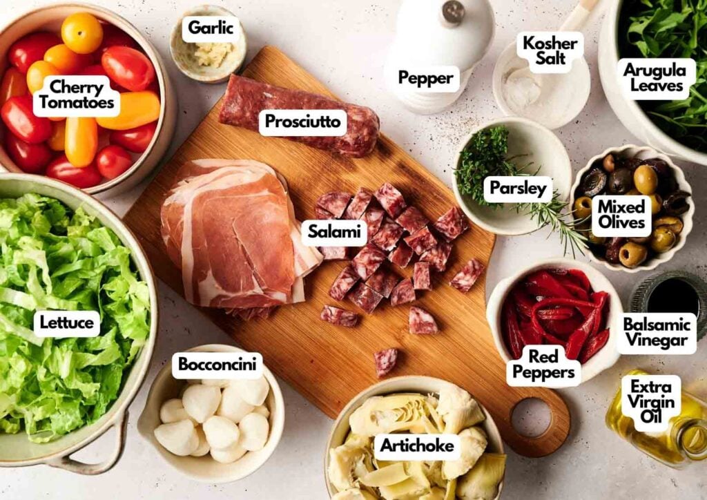 A wooden board with prosciutto and salami, surrounded by labeled bowls of lettuce, cherry tomatoes, bocconcini, artichoke, garlic, olives, arugula&mdash;everything you need to create a vibrant antipasto salad&mdash;plus parsley, red peppers, vinegar, oil, salt, and pepper.