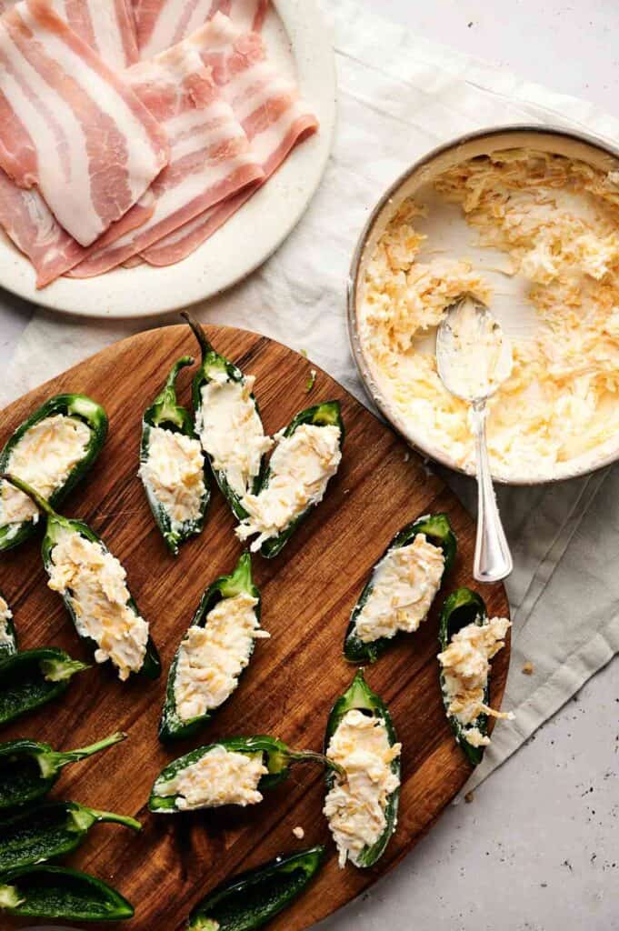 Jalapeño halves filled with a creamy cheese mixture rest on a wooden board, while sliced bacon awaits to embrace them, creating irresistible bacon-wrapped jalapeño poppers. A bowl of leftover cheese mixture sits enticingly beside them.
