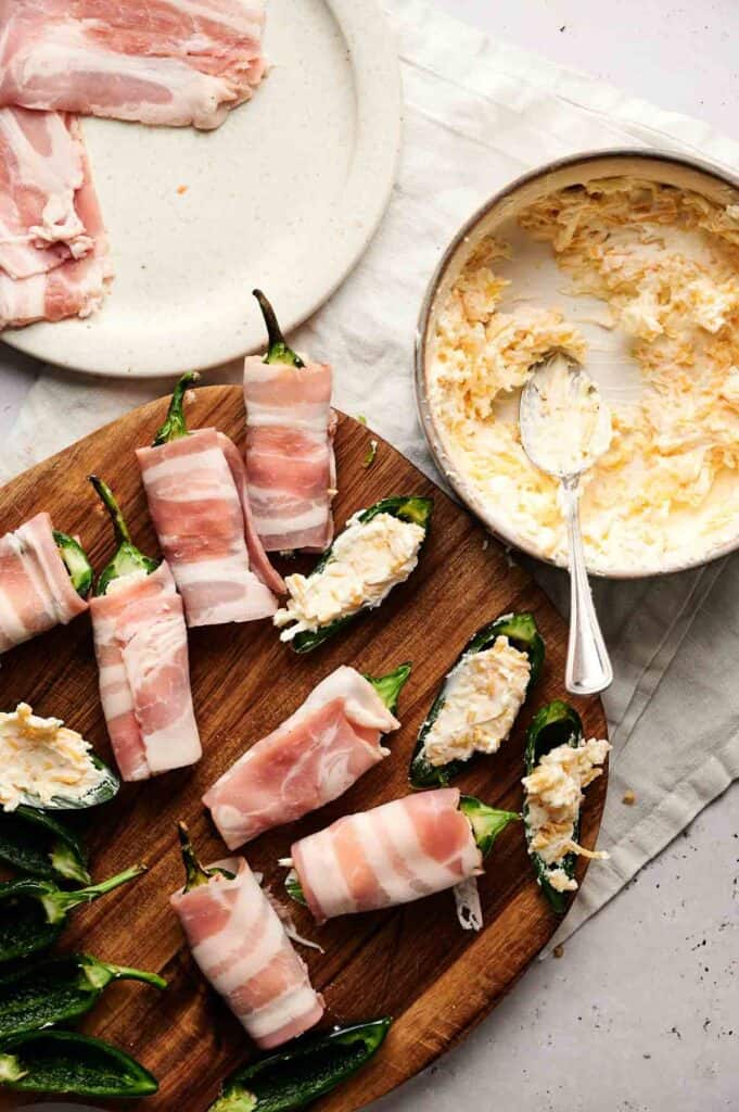Bacon-wrapped jalape&ntilde;o poppers sit enticingly on a wooden board, with halves filled to the brim with cheese. Next to them, a bowl of cheese mixture awaits alongside crispy slices of bacon, ready to add an irresistible twist.