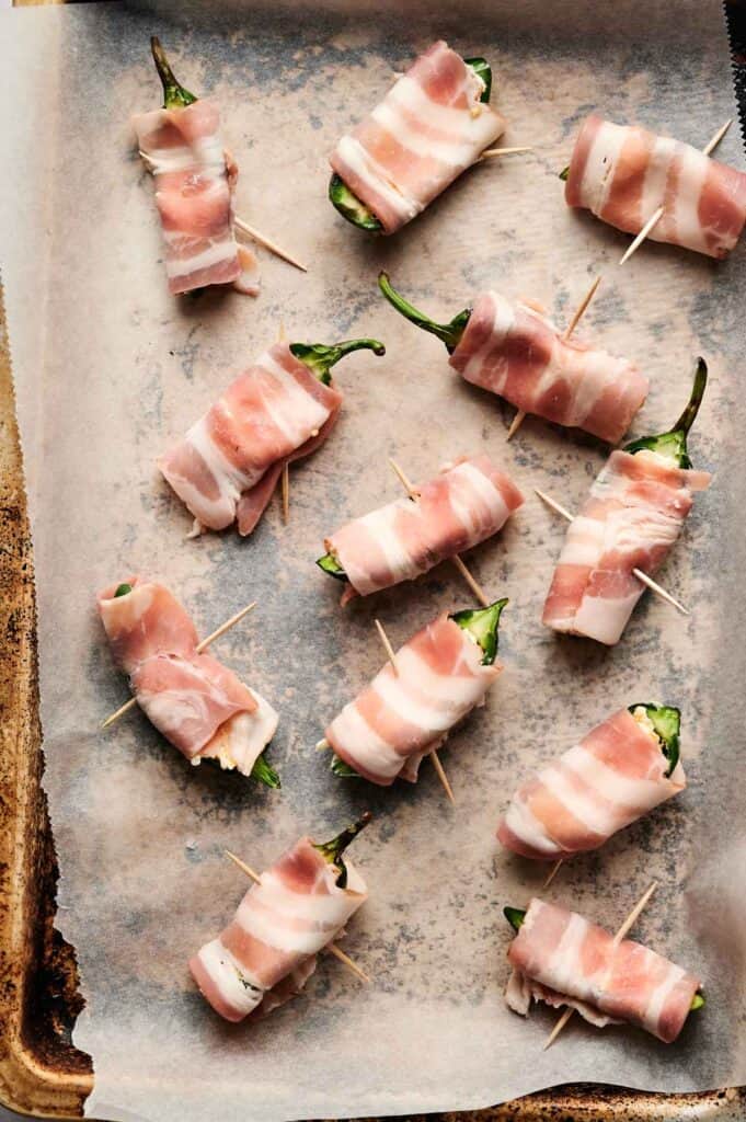 Bacon-wrapped jalapeño poppers rest on a parchment-lined baking sheet, each secured with toothpicks.
