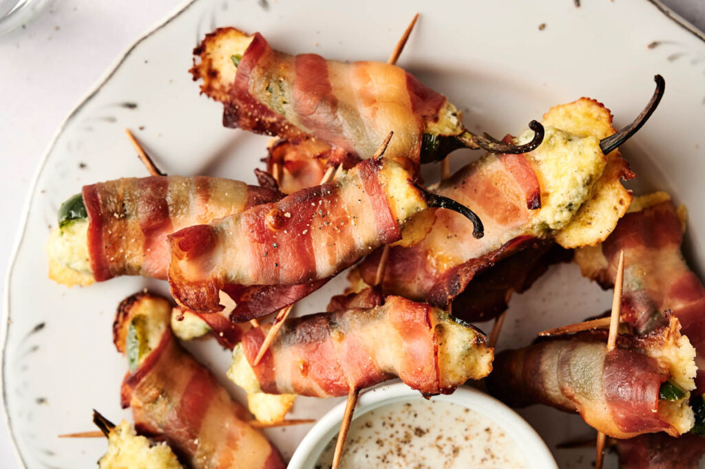 Bacon wrapped jalapeno poppers with toothpicks rest on a white plate, served alongside a bowl of creamy dipping sauce.
